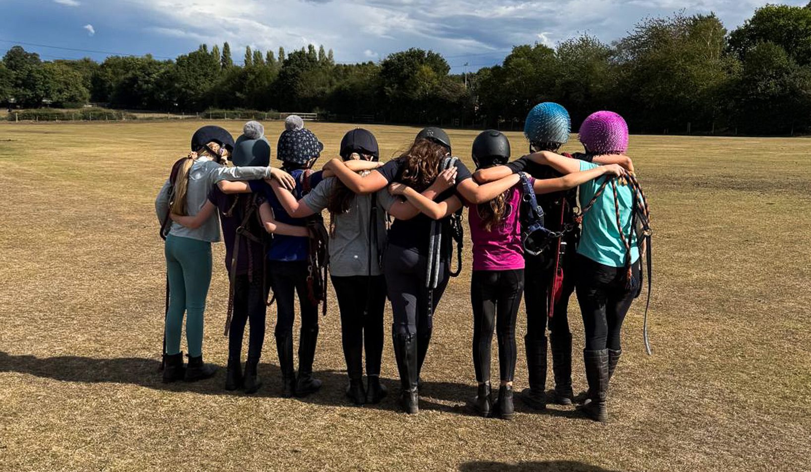 A group of children stand in a field with their backs to their camera and arms on each others shoulders during a holiday activity at Bourne Vale Stables