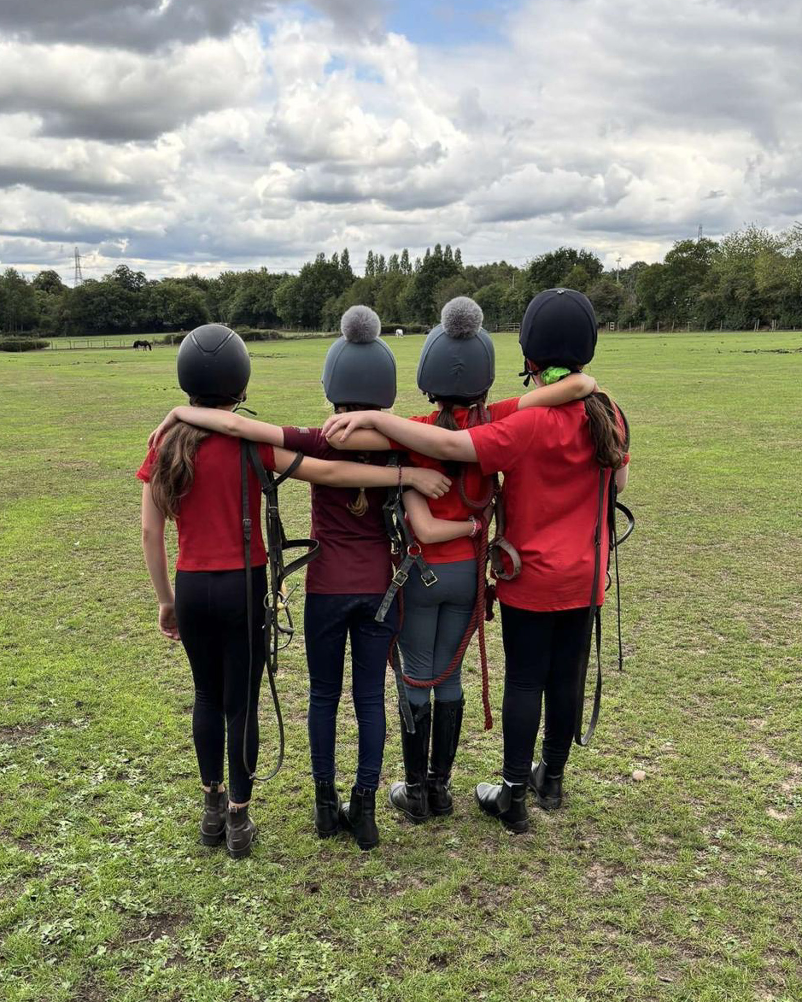 4 young riders stand arm in arm with their backs to the camera, looking out over the field
